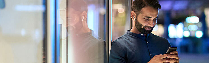 man leaning against window with earpods in and smiling at his phone at night